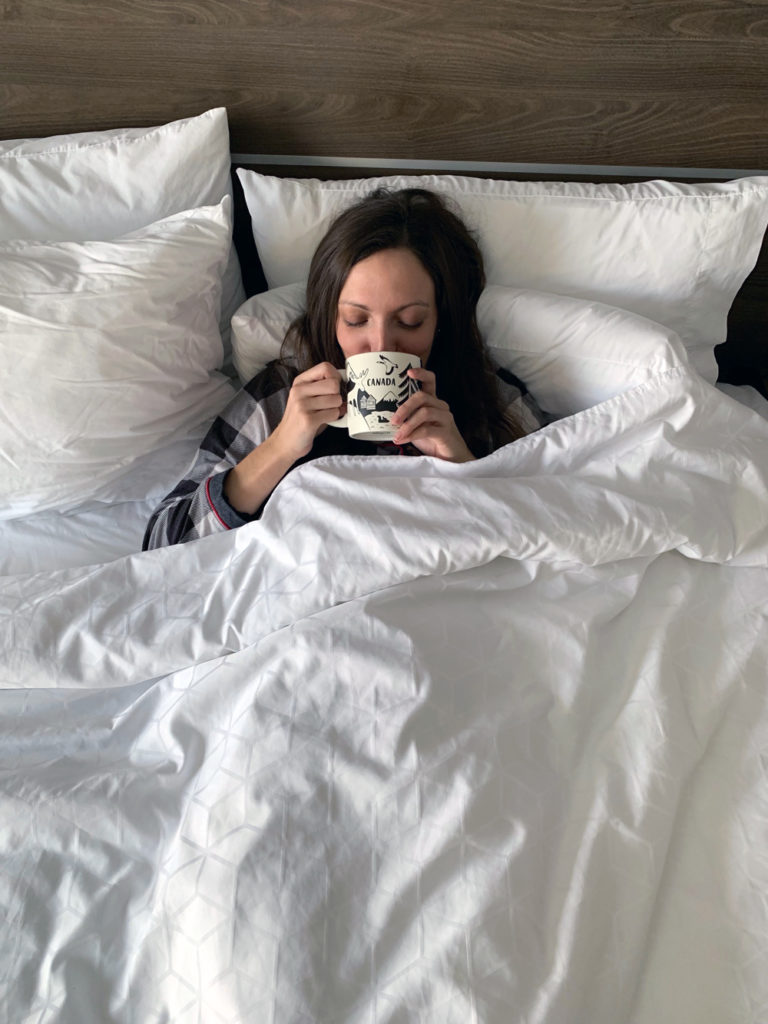 Lauren laying in a hotel bed sipping coffee.