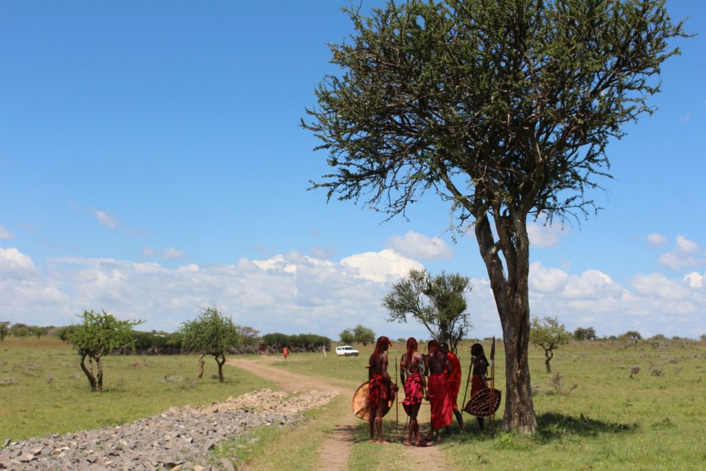 Members of the Maasai Mara tribe standing in a field next to a tree catching shade.