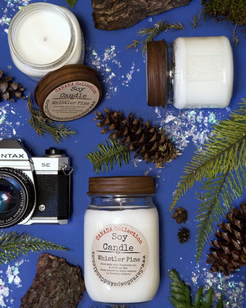 Shop local with Sara's Soaps & Candles. Candle laying on blue surface with trees and nature props surrounding it.